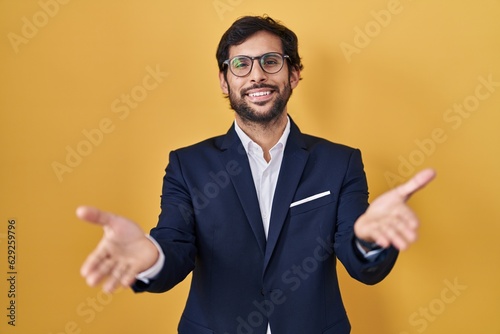 Handsome latin man standing over yellow background smiling cheerful offering hands giving assistance and acceptance.