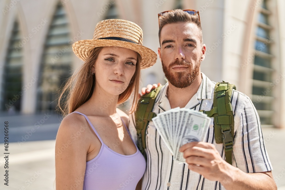 Young tourist couple holding dollars banknotes thinking attitude and sober expression looking self confident