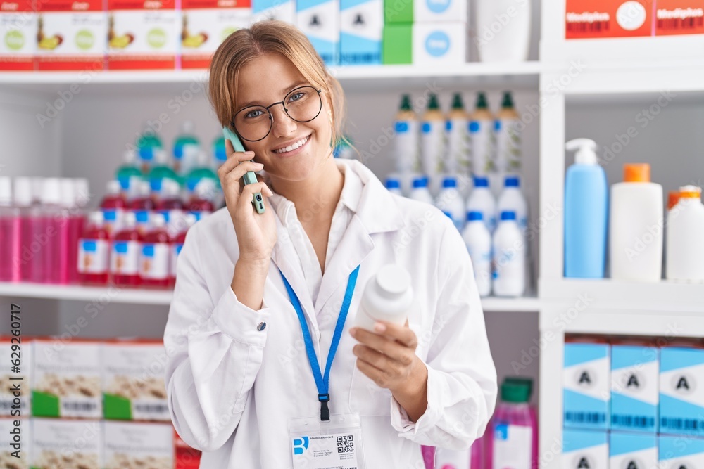 Young blonde girl pharmacist holding pills bottle talking on smartphone at pharmacy