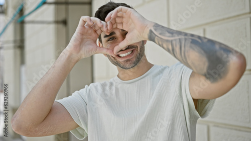 Young hispanic man smiling confident doing heart gesture with hands at street