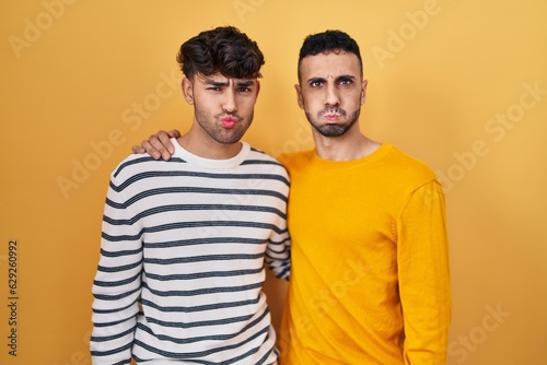 Young hispanic gay couple standing over yellow background puffing cheeks with funny face. mouth inflated with air  crazy expression.