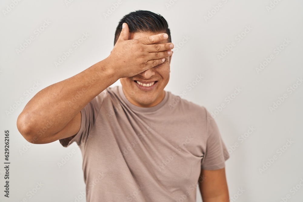 Hispanic young man standing over white background smiling and laughing with hand on face covering eyes for surprise. blind concept. Hispanic young man standing over white background smiling and laughing with hand on face covering eyes for surprise. blind concept.
