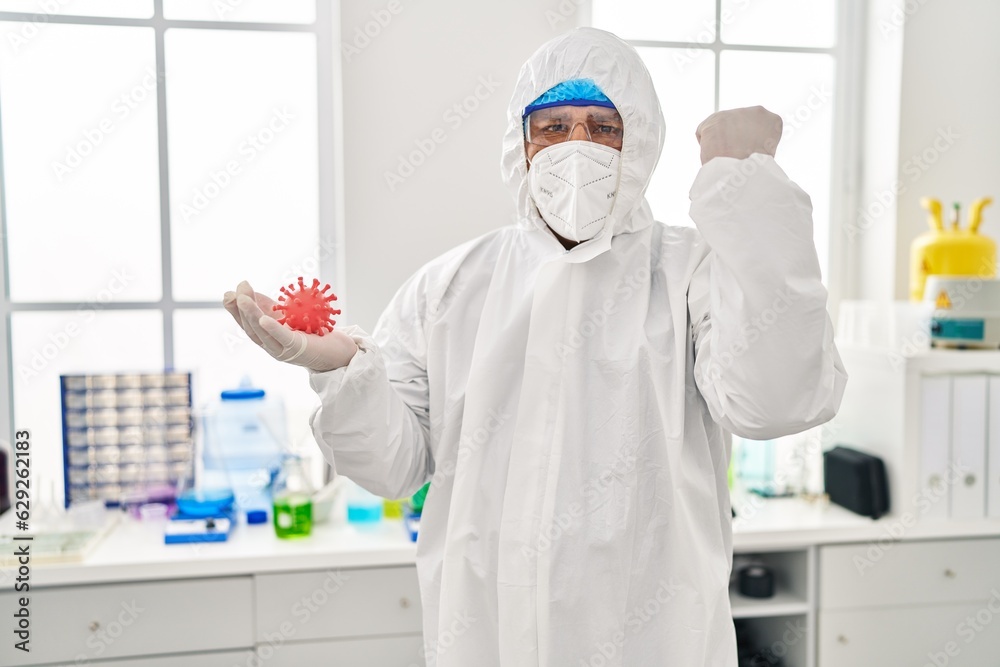 Hispanic young man working at scientist laboratory holding virus toy annoyed and frustrated shouting with anger, yelling crazy with anger and hand raised Hispanic young man working at scientist laboratory holding virus toy annoyed and frustrated shouting with anger, yelling crazy with anger and hand raised