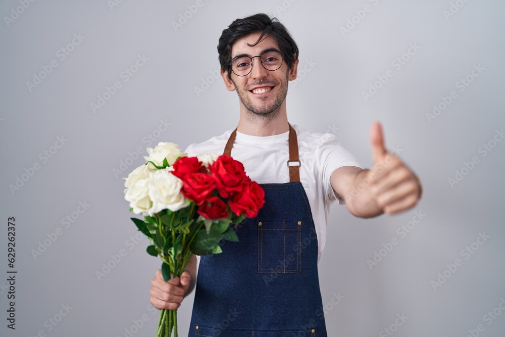Young hispanic man holding bouquet of white and red roses approving doing positive gesture with hand, thumbs up smiling and happy for success. winner gesture. Young hispanic man holding bouquet of white and red roses approving doing positive gesture with hand, thumbs up smiling and happy for success. winner gesture.