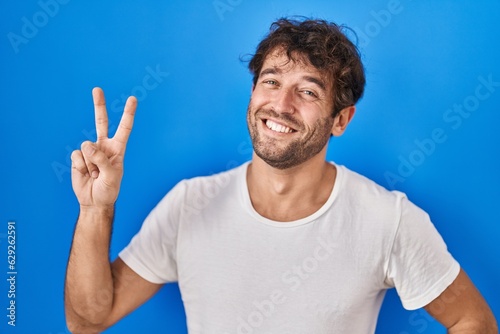 Hispanic young man standing over blue background smiling looking to the camera showing fingers doing victory sign. number two.