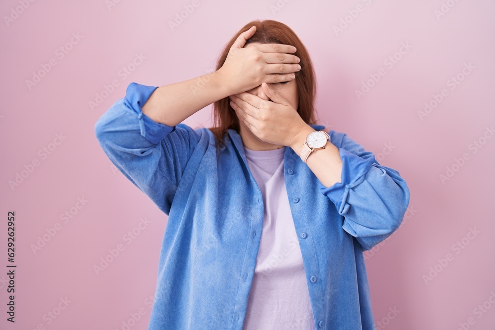 Young hispanic woman with red hair standing over pink background covering eyes and mouth with hands, surprised and shocked. hiding emotion Young hispanic woman with red hair standing over pink background covering eyes and mouth with hands, surprised and shocked. hiding emotion