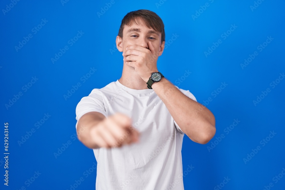 Caucasian blond man standing over blue background laughing at you, pointing finger to the camera with hand over mouth, shame expression Caucasian blond man standing over blue background laughing at you, pointing finger to the camera with hand over mouth, shame expression