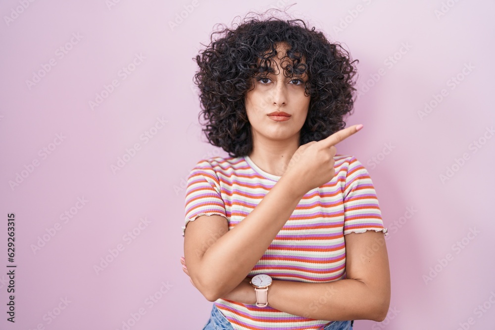 Young middle east woman standing over pink background pointing with hand finger to the side showing advertisement, serious and calm face Young middle east woman standing over pink background pointing with hand finger to the side showing advertisement, serious and calm face