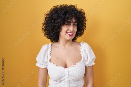 Young brunette woman with curly hair standing over yellow background winking looking at the camera with sexy expression, cheerful and happy face. © Krakenimages.com