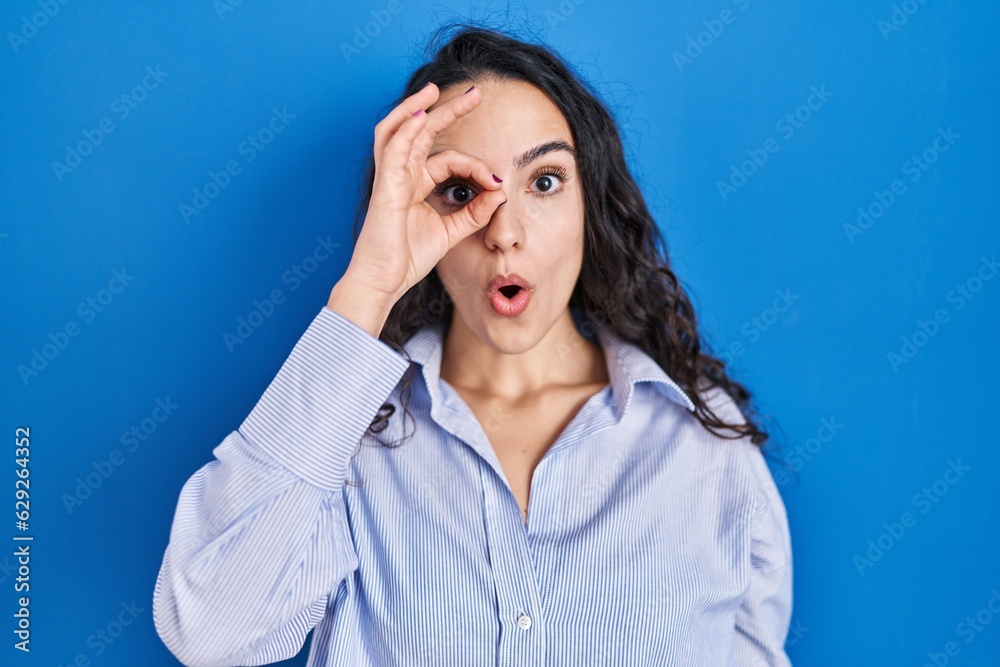 Young brunette woman standing over blue background doing ok gesture shocked with surprised face, eye looking through fingers. unbelieving expression. Young brunette woman standing over blue background doing ok gesture shocked with surprised face, eye looking through fingers. unbelieving expression.