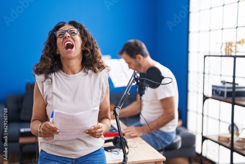 Middle age hispanic woman singing at music studio angry and mad screaming frustrated and furious, shouting with anger looking up.