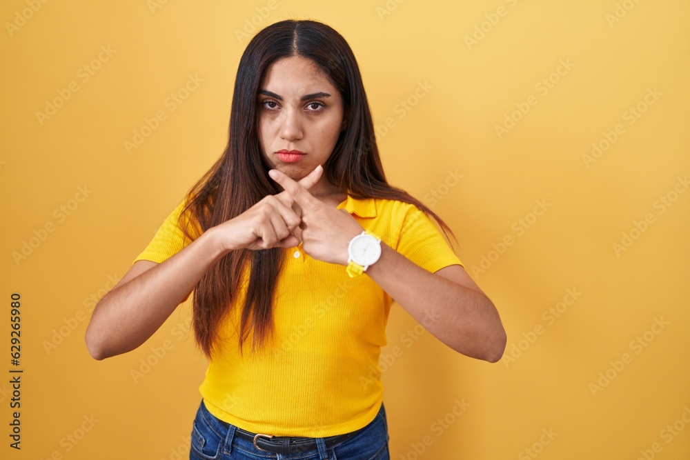 Young arab woman standing over yellow background rejection expression crossing fingers doing negative sign Young arab woman standing over yellow background rejection expression crossing fingers doing negative sign