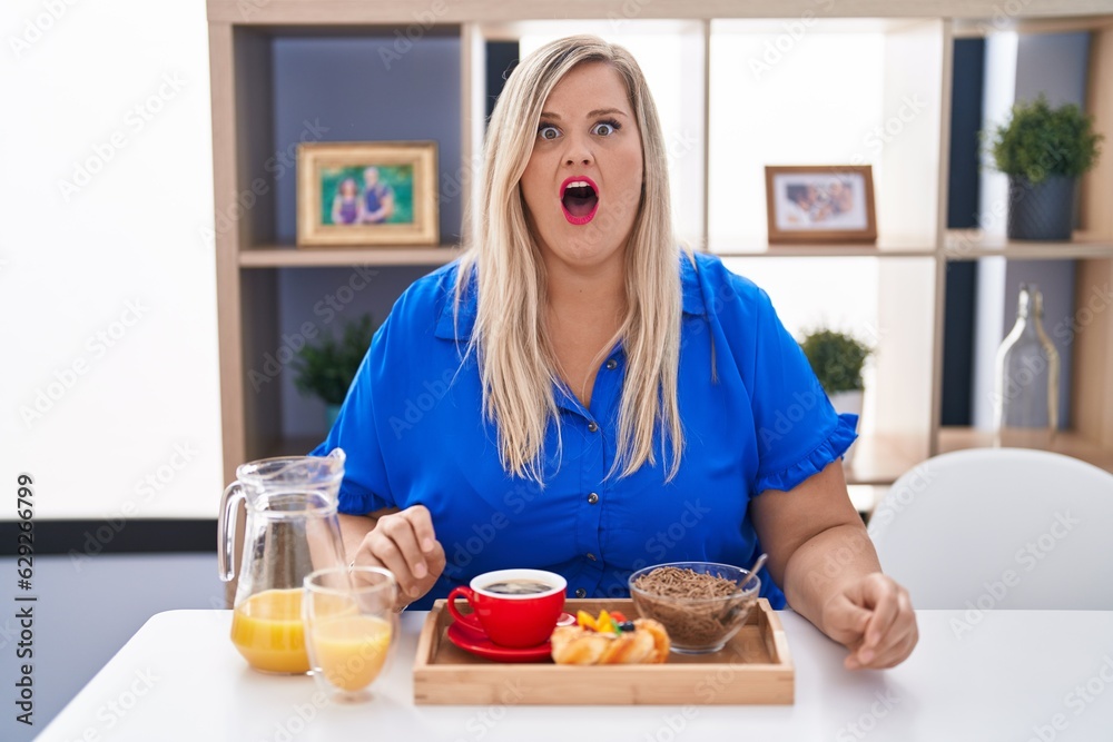 Caucasian plus size woman eating breakfast at home afraid and shocked with surprise expression, fear and excited face.
