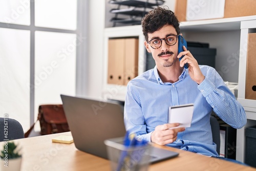 Young caucasian man business worker talking on smartphone reading notebook at office
