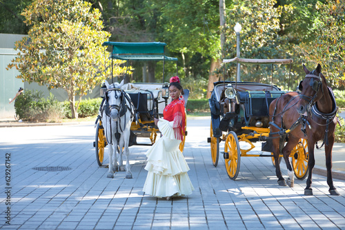 Photography Young black and South American woman wearing a beige gypsy flamenco suit and red shawl, posing next to typical horse-drawn carriages of the city of Seville in Spain