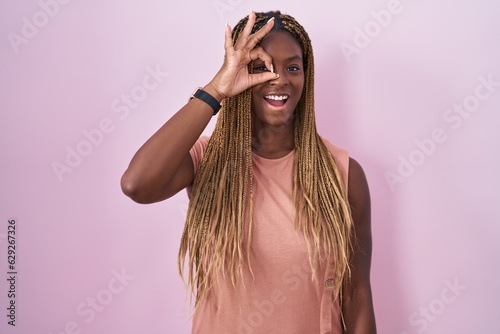 African american woman with braided hair standing over pink background doing ok gesture with hand smiling, eye looking through fingers with happy face. © Krakenimages.com