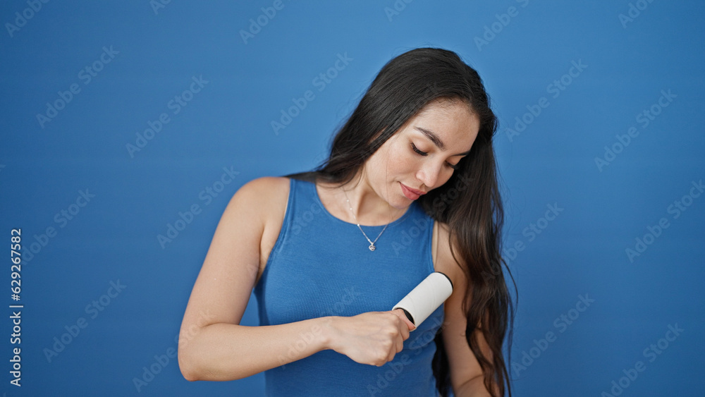 Young beautiful hispanic woman cleaning t shirt with pet hair roller over isolated blue background Young beautiful hispanic woman cleaning t shirt with pet hair roller over isolated blue background