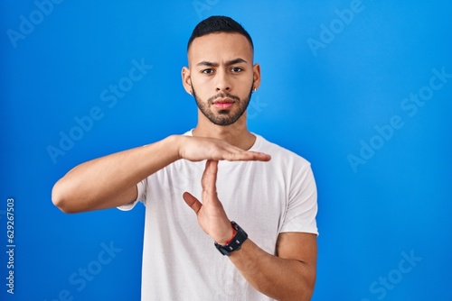 Young hispanic man standing over blue background doing time out gesture with hands, frustrated and serious face