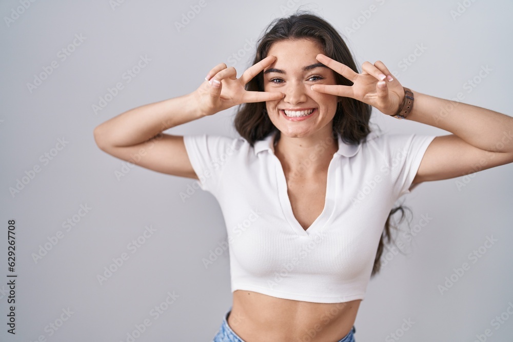 Young teenager girl standing over white background doing peace symbol with fingers over face, smiling cheerful showing victory Young teenager girl standing over white background doing peace symbol with fingers over face, smiling cheerful showing victory