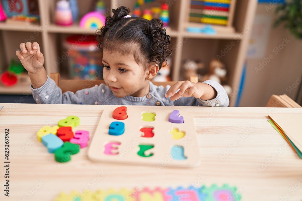 Fototapeta premium Adorable hispanic girl playing with maths puzzle game sitting on table at kindergarten