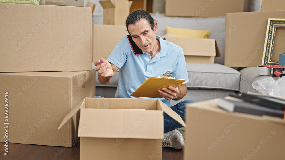 Middle age man checking cardboard box writing on clipboard talking on smartphone at new home Middle age man checking cardboard box writing on clipboard talking on smartphone at new home