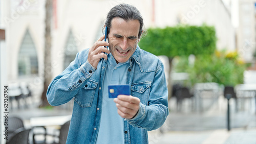Middle age man holding credit card arguing by smartphone at coffee shop terrace