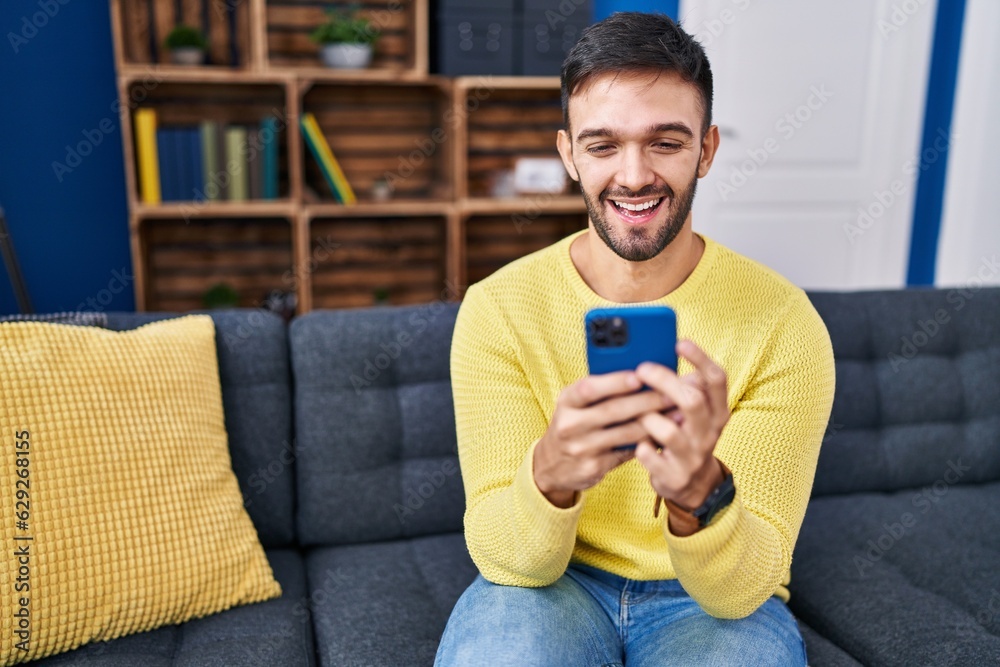 Young hispanic man using smartphone sitting on sofa at home