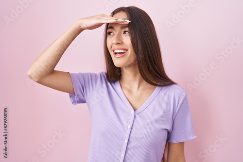 Young hispanic woman with long hair standing over pink background very happy and smiling looking far away with hand over head. searching concept.