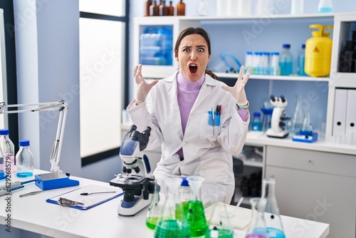 Young brunette woman working at scientist laboratory crazy and mad shouting and yelling with aggressive expression and arms raised. frustration concept.