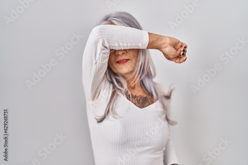Middle age woman with grey hair standing over white background covering eyes with arm smiling cheerful and funny. blind concept.