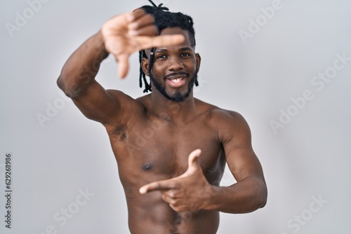 African man with dreadlocks standing shirtless over isolated background smiling making frame with hands and fingers with happy face. creativity and photography concept.
