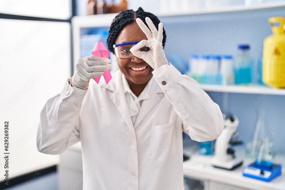 African american woman working at scientist laboratory holding pink ribbon smiling happy doing ok sign with hand on eye looking through fingers