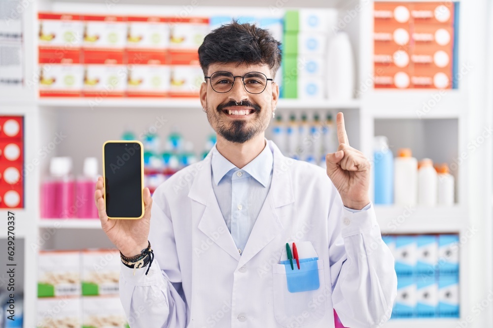 Young hispanic man with beard working at pharmacy drugstore showing smartphone screen smiling with an idea or question pointing finger with happy face, number one Young hispanic man with beard working at pharmacy drugstore showing smartphone screen smiling with an idea or question pointing finger with happy face, number one