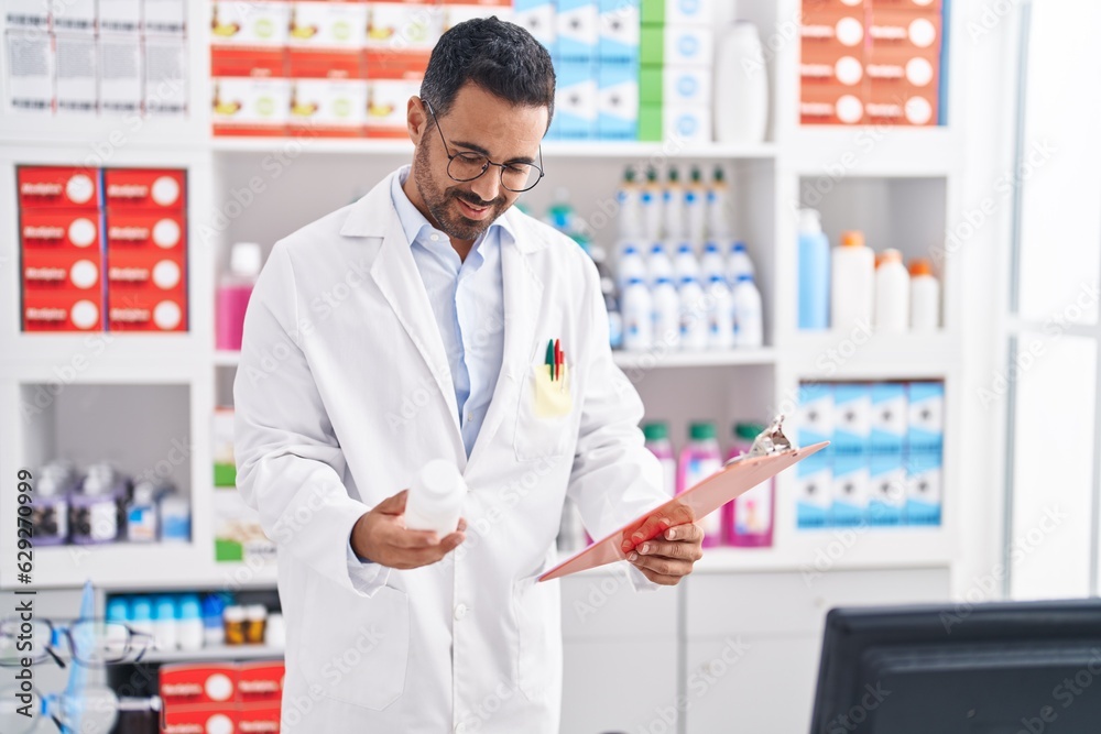 Young hispanic man pharmacist holding pills bottle reading document at pharmacy