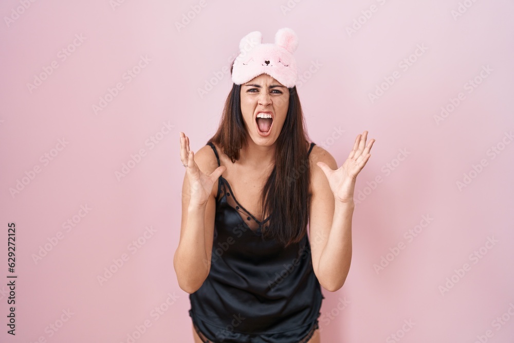 Young brunette woman wearing sleep mask and pyjama crazy and mad shouting and yelling with aggressive expression and arms raised. frustration concept. Young brunette woman wearing sleep mask and pyjama crazy and mad shouting and yelling with aggressive expression and arms raised. frustration concept.