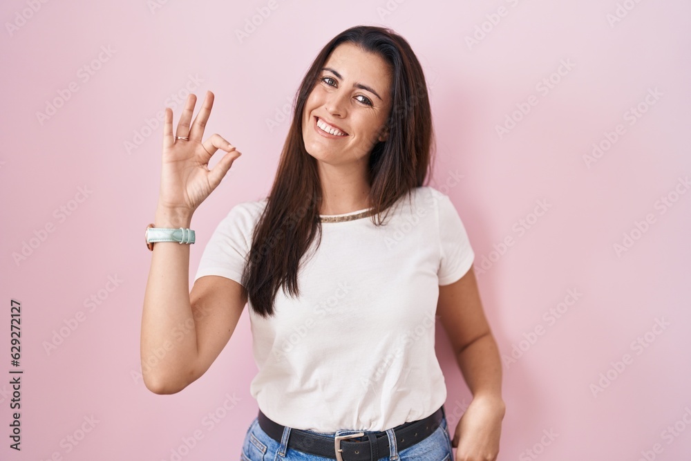 Young brunette woman standing over pink background smiling positive doing ok sign with hand and fingers. successful expression. Young brunette woman standing over pink background smiling positive doing ok sign with hand and fingers. successful expression.