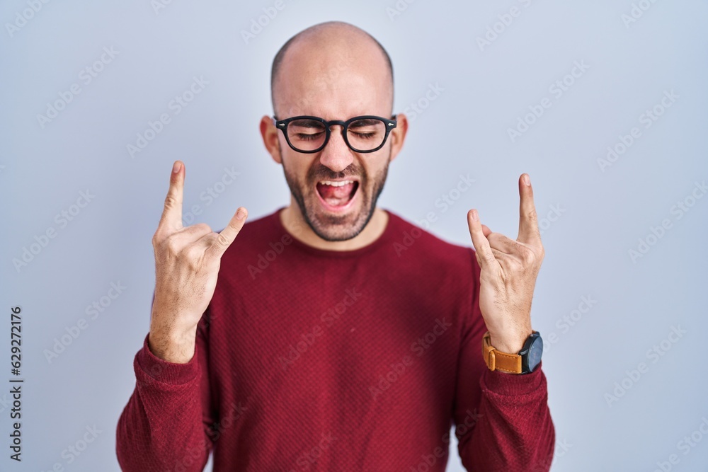Young bald man with beard standing over white background wearing glasses shouting with crazy expression doing rock symbol with hands up. music star. heavy music concept. Young bald man with beard standing over white background wearing glasses shouting with crazy expression doing rock symbol with hands up. music star. heavy music concept.