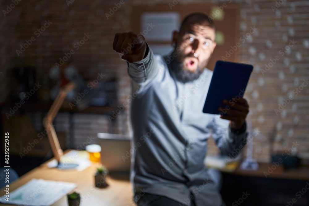 Young hispanic man with beard and tattoos working at the office at night pointing with finger surprised ahead, open mouth amazed expression, something on the front Young hispanic man with beard and tattoos working at the office at night pointing with finger surprised ahead, open mouth amazed expression, something on the front