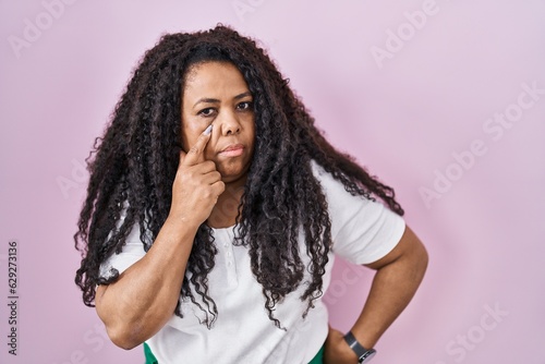 Plus size hispanic woman standing over pink background pointing to the eye watching you gesture, suspicious expression