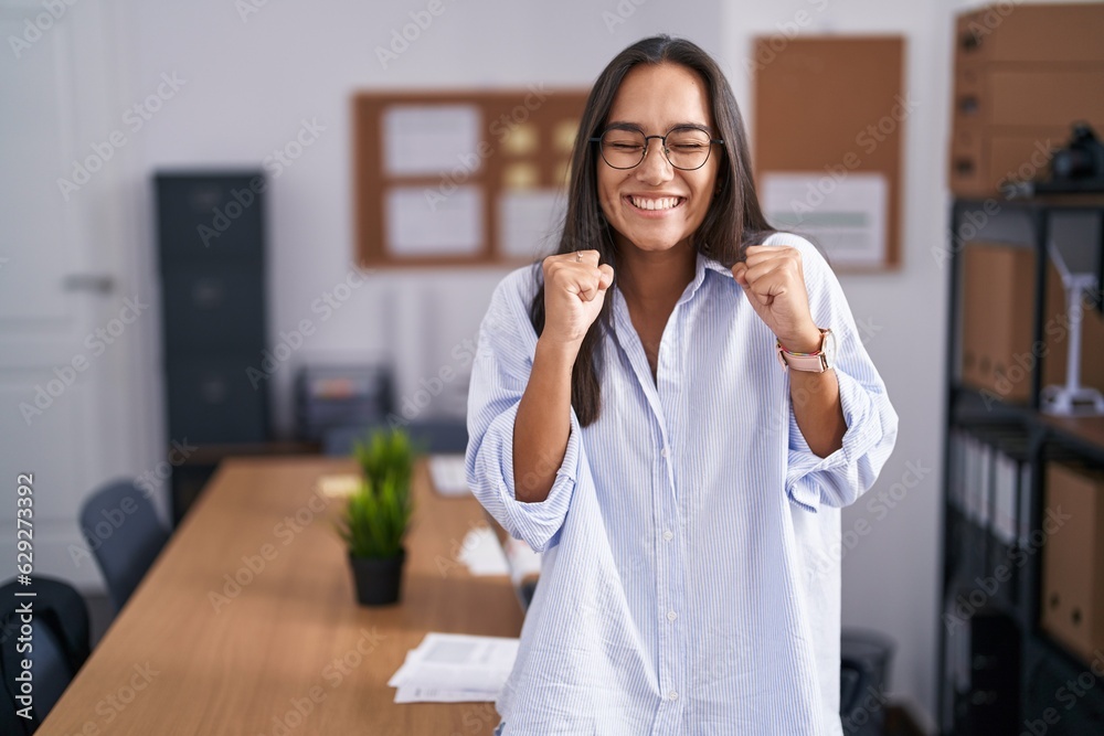 Young hispanic woman at the office excited for success with arms raised and eyes closed celebrating victory smiling. winner concept. Young hispanic woman at the office excited for success with arms raised and eyes closed celebrating victory smiling. winner concept.
