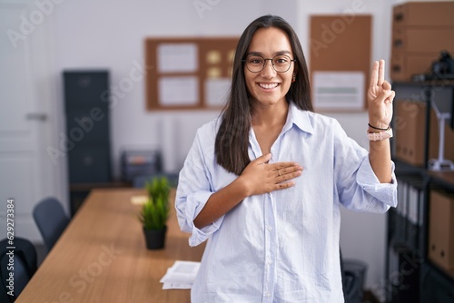 Young hispanic woman at the office smiling swearing with hand on chest and fingers up, making a loyalty promise oath