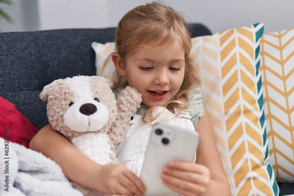 Adorable blonde girl using smartphone hugging teddy bear at home Adorable blonde girl using smartphone hugging teddy bear at home