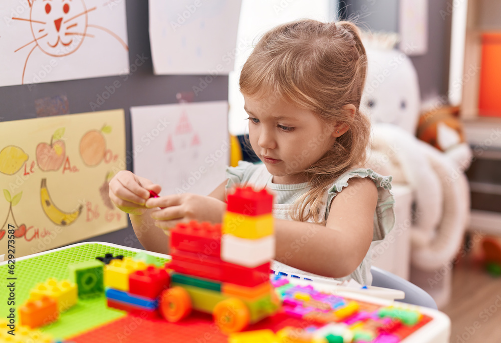 Adorable blonde girl playing with construction blocks sitting on table at kindergarten Adorable blonde girl playing with construction blocks sitting on table at kindergarten