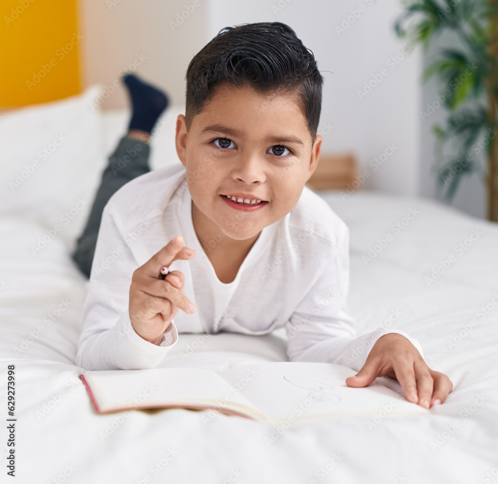 Adorable hispanic boy writing on notebook lying on bed at bedroom Adorable hispanic boy writing on notebook lying on bed at bedroom