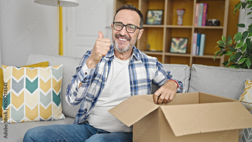 Middle age man unpacking cardboard box with thumb up gesture at home Middle age man unpacking cardboard box with thumb up gesture at home