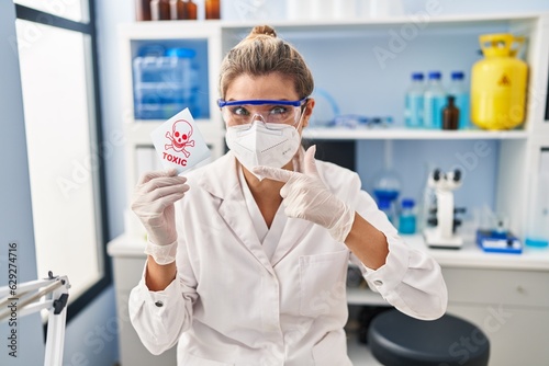 Young woman working at scientist laboratory holding toxic banner smiling happy pointing with hand and finger