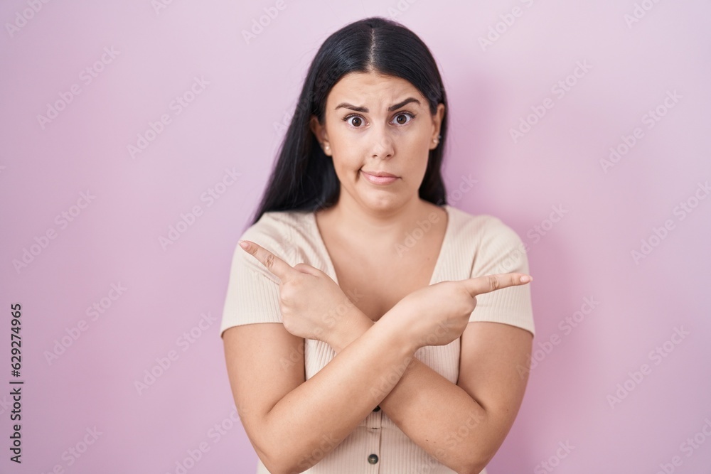 Young hispanic woman standing over pink background pointing to both sides with fingers, different direction disagree