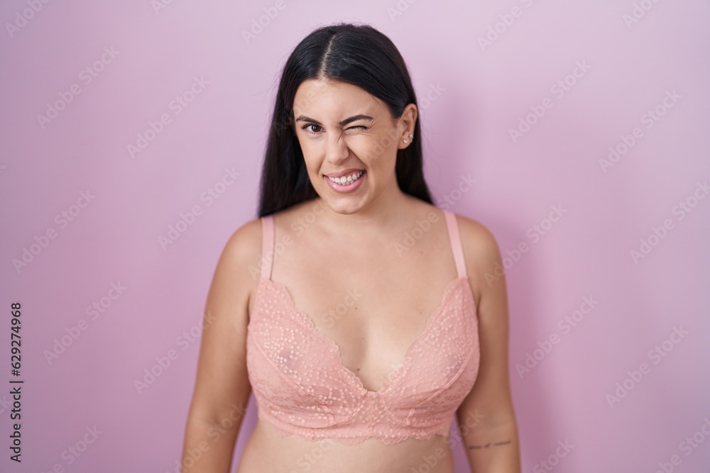 Young hispanic woman wearing pink bra winking looking at the camera with sexy expression, cheerful and happy face.