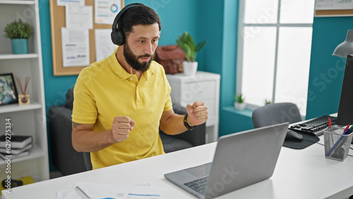 Young hispanic man business worker using laptop and headphones celebrating at the office