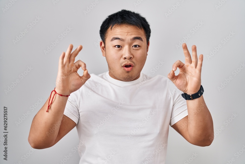 Fototapeta premium Young chinese man standing over white background looking surprised and shocked doing ok approval symbol with fingers. crazy expression